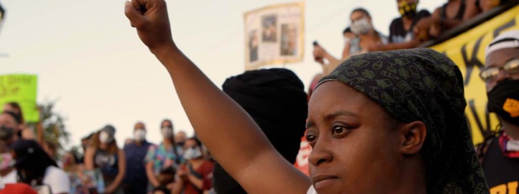 Black woman wearing a head wrap with her fist in the air at a demonstration.