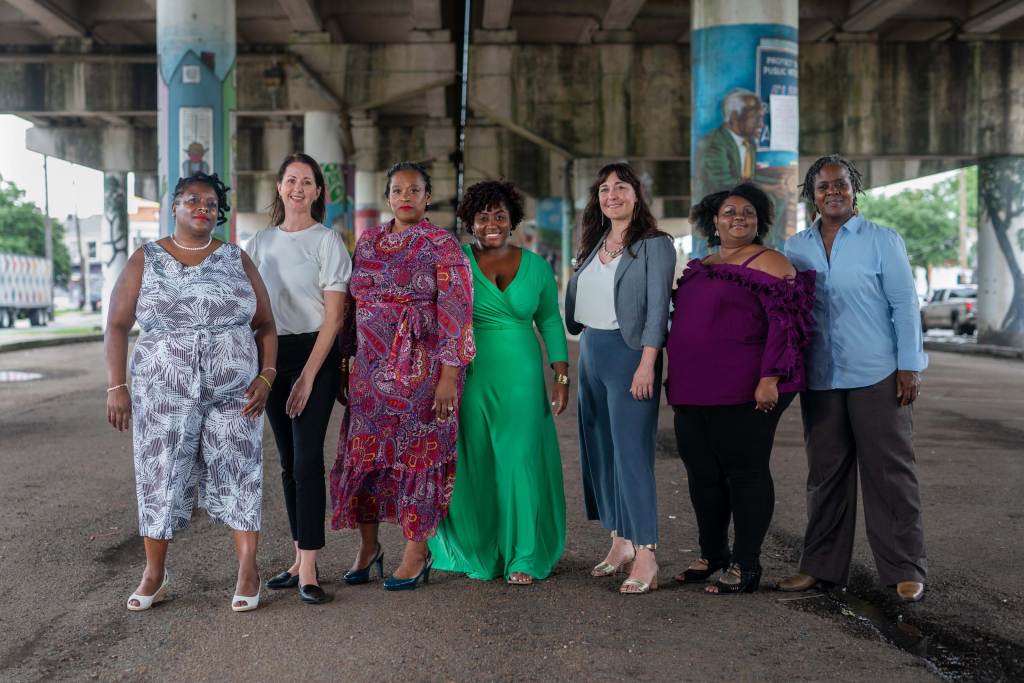 Eight women standing together and smiling in front of a highway overpass. Columns behind them are painted with unique designs depicting significant people and moments in New Orleans history.