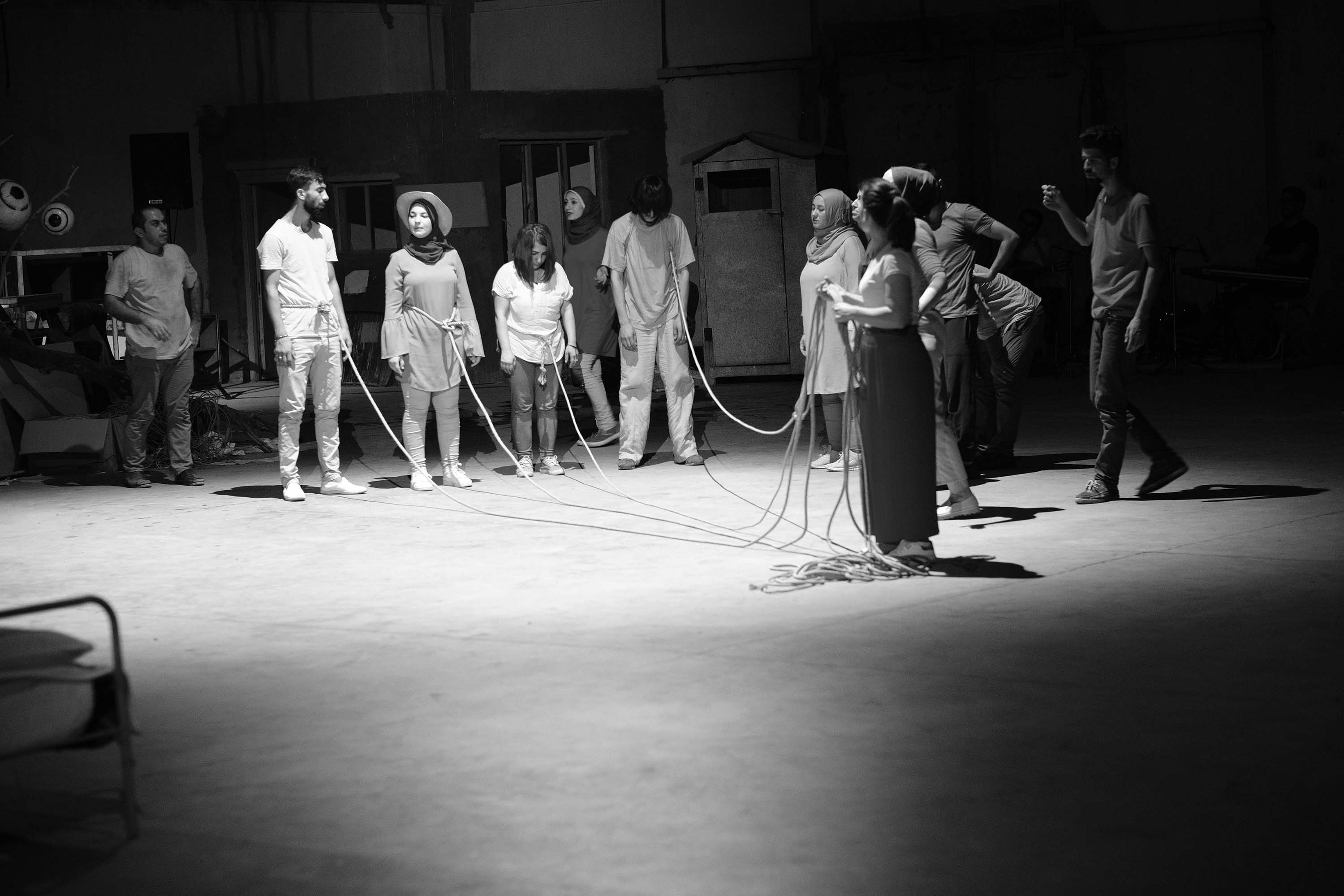  A series of black-and-whie images showing actors on a stage while moving around, holding hands and bowing in a line, standing in two lines across from each other holding ropes and in a single line dancing.