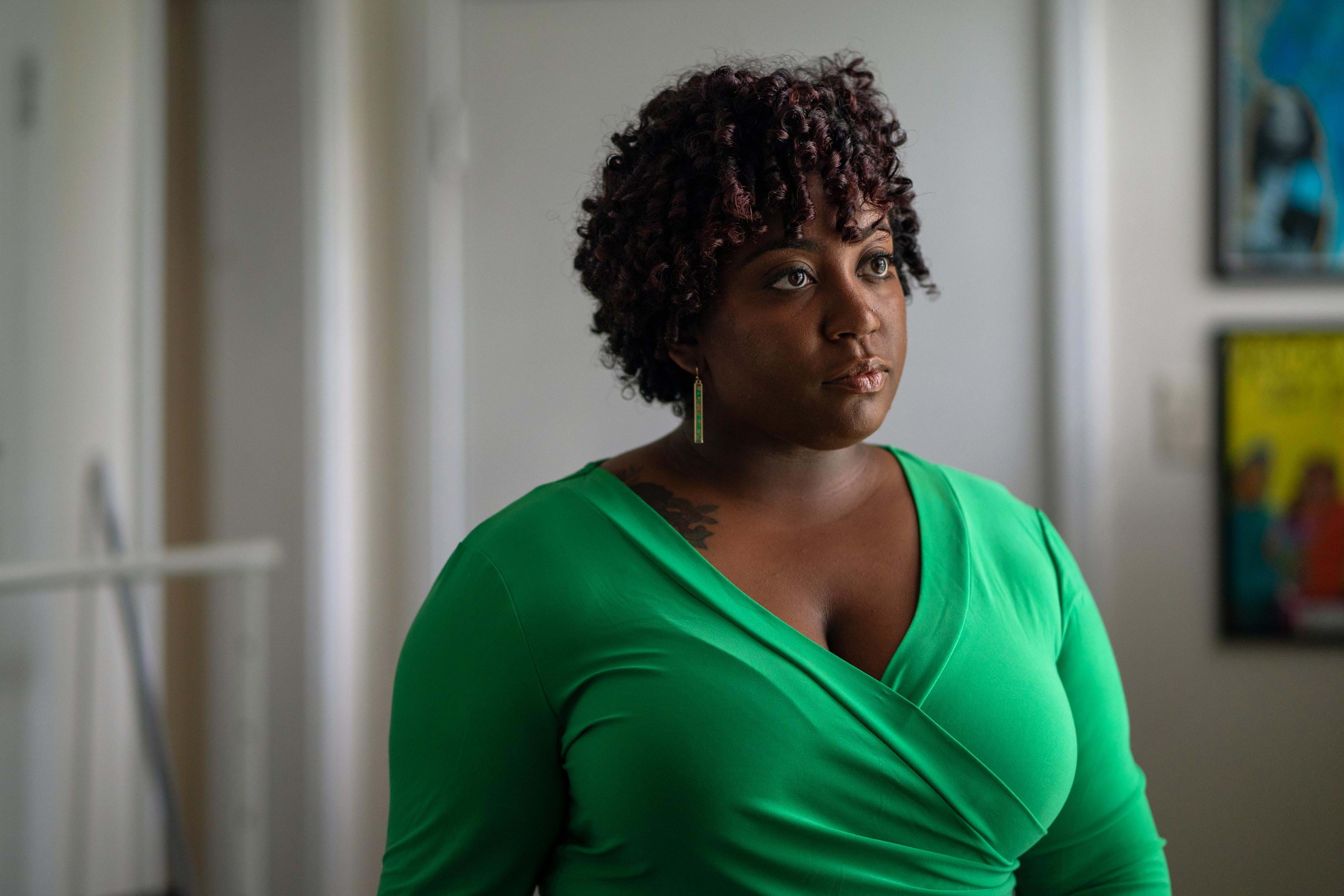 Elyse, a Black woman, wearing a bright green wrap dress gazes softly in an office decorated with posters.