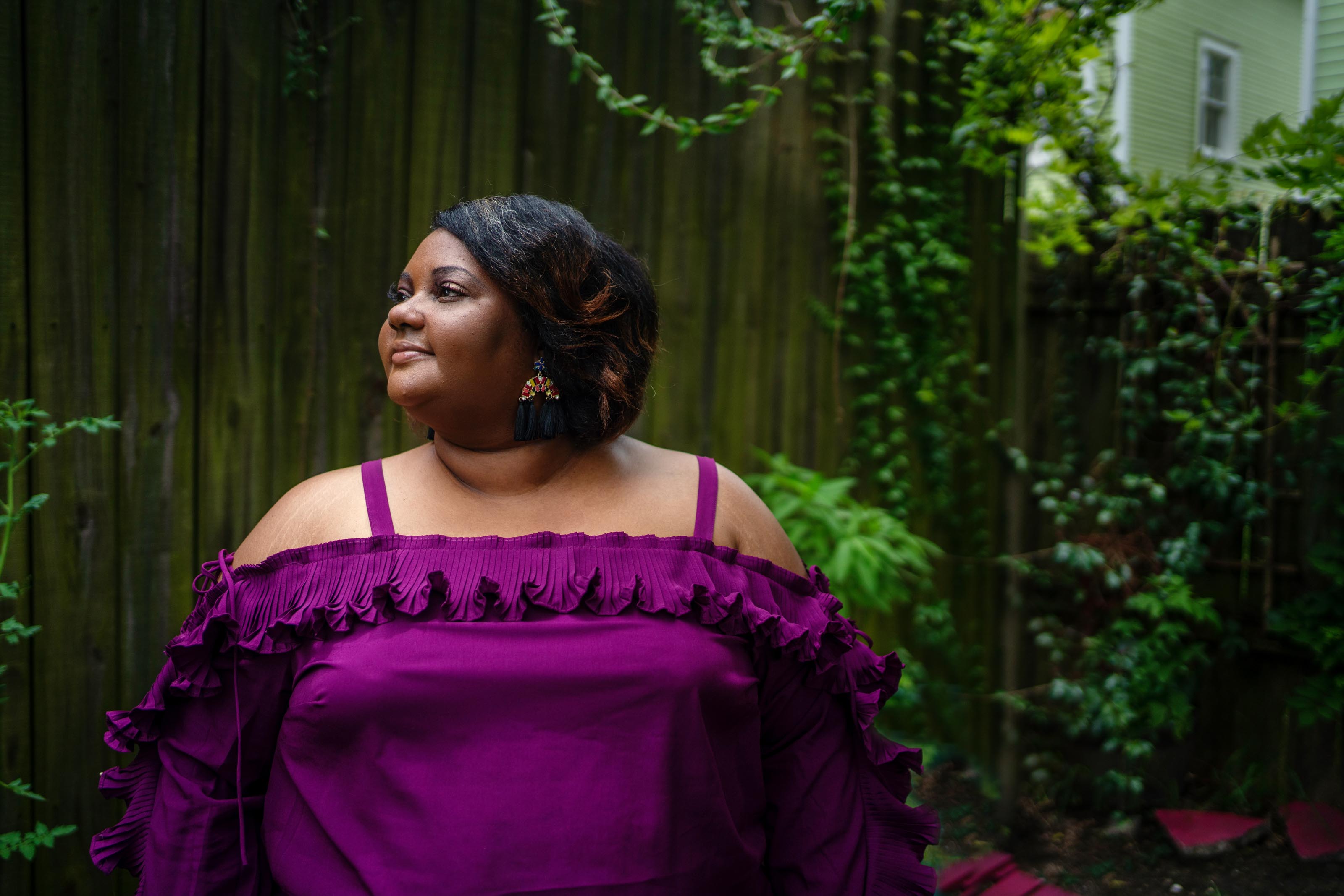 Raven, a Black woman, holds her head high, wearing a purple ruffled top with a fence behind her decorated with plants and ivy.