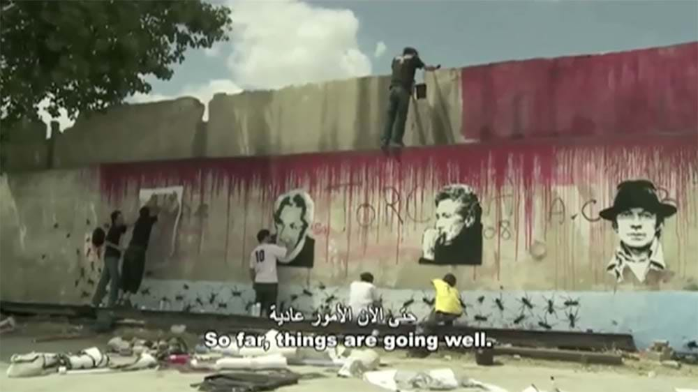 A wide shot of a wall with a mural being painted. Red paint drips down onto black and white faces and people spray paint along the wall.