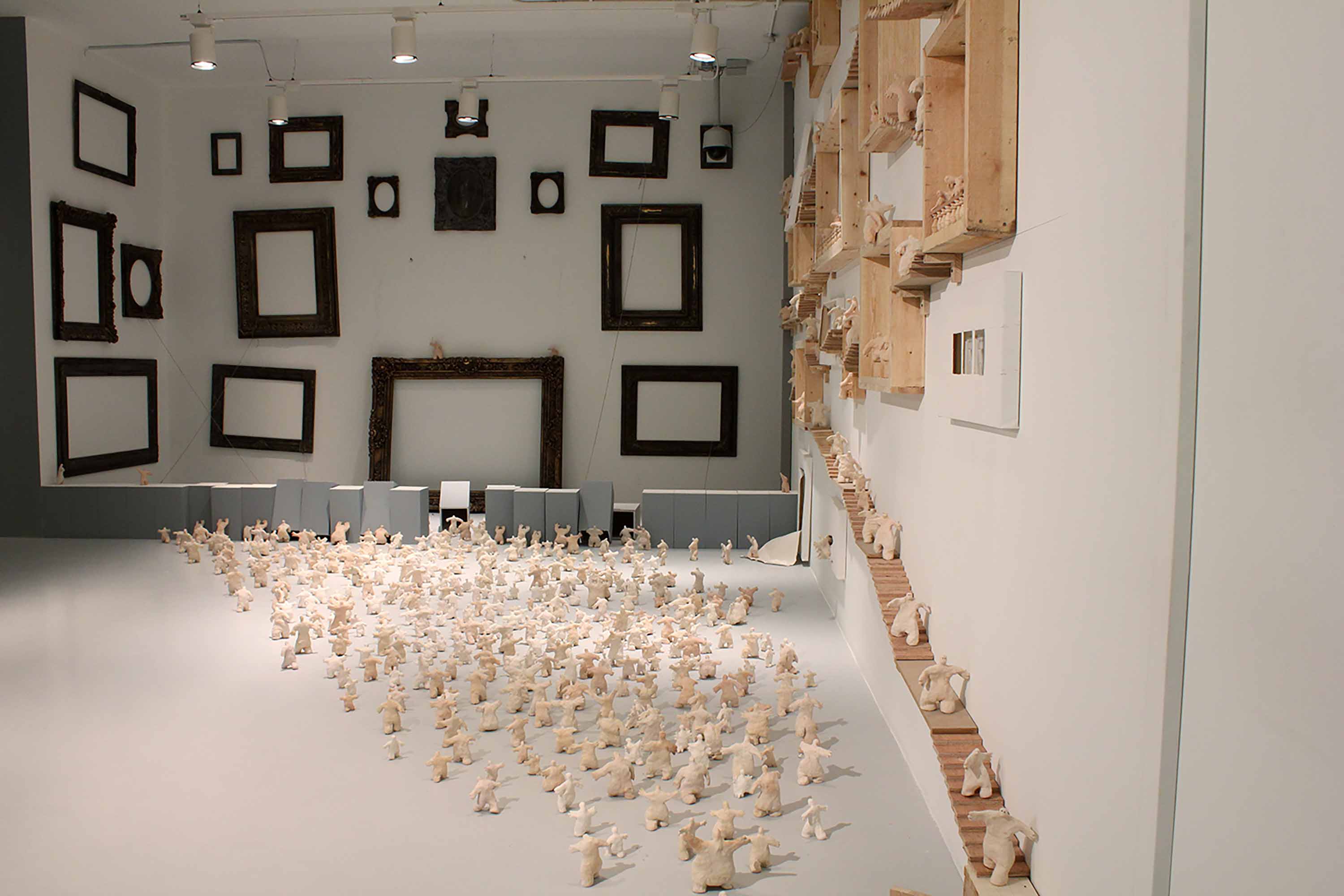 A gallery space with one corner covered in small white figurines on the floor and climbing the wall, knocking down columns and taking over a wooden staircase and windows on the wall.