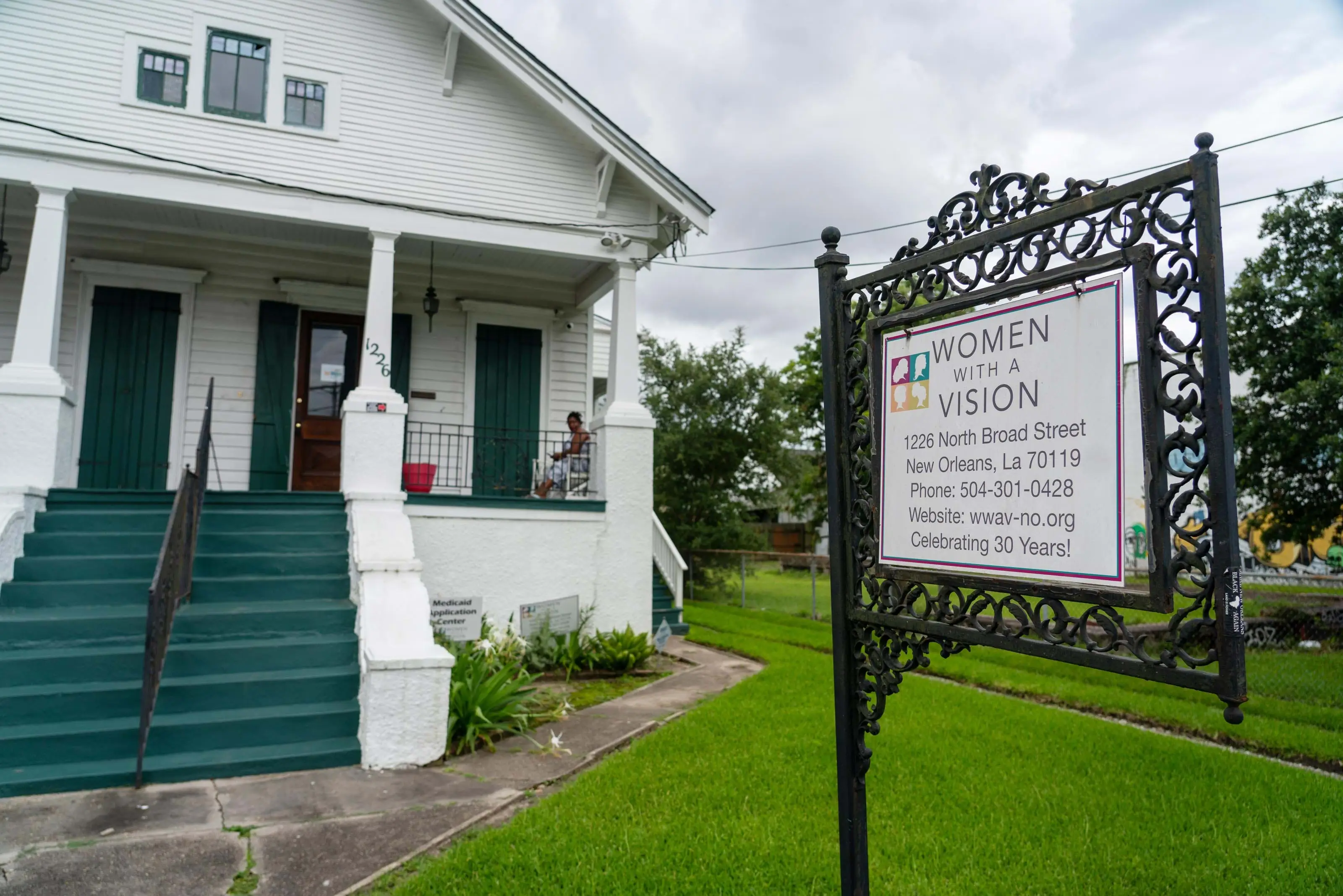 A white Bungalow house with a flagstone walkway and manicured lawn. A sign hanging from a wrought iron post reads, "Women with a Vision”