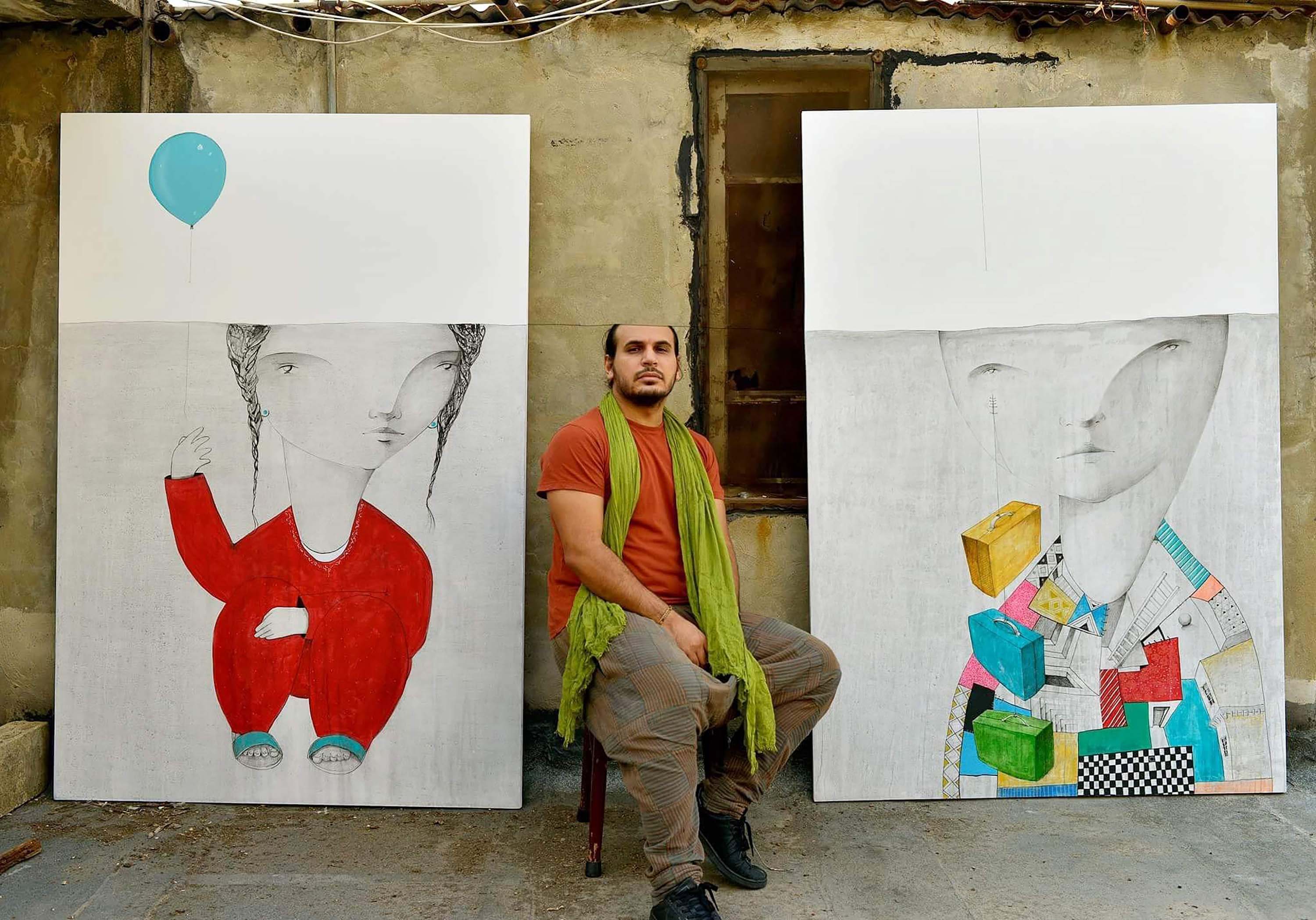 Mohamad Kahyata sits between two of his works of art on large white canvases. He is wearing an orange shirt and green scarf and has a short beard. The artworks depict a sketched figure on each, with oversized heads and some painted color on their bodies. Their heads cut off along the same line as Khayata’s, creating a straight line across all three heads.