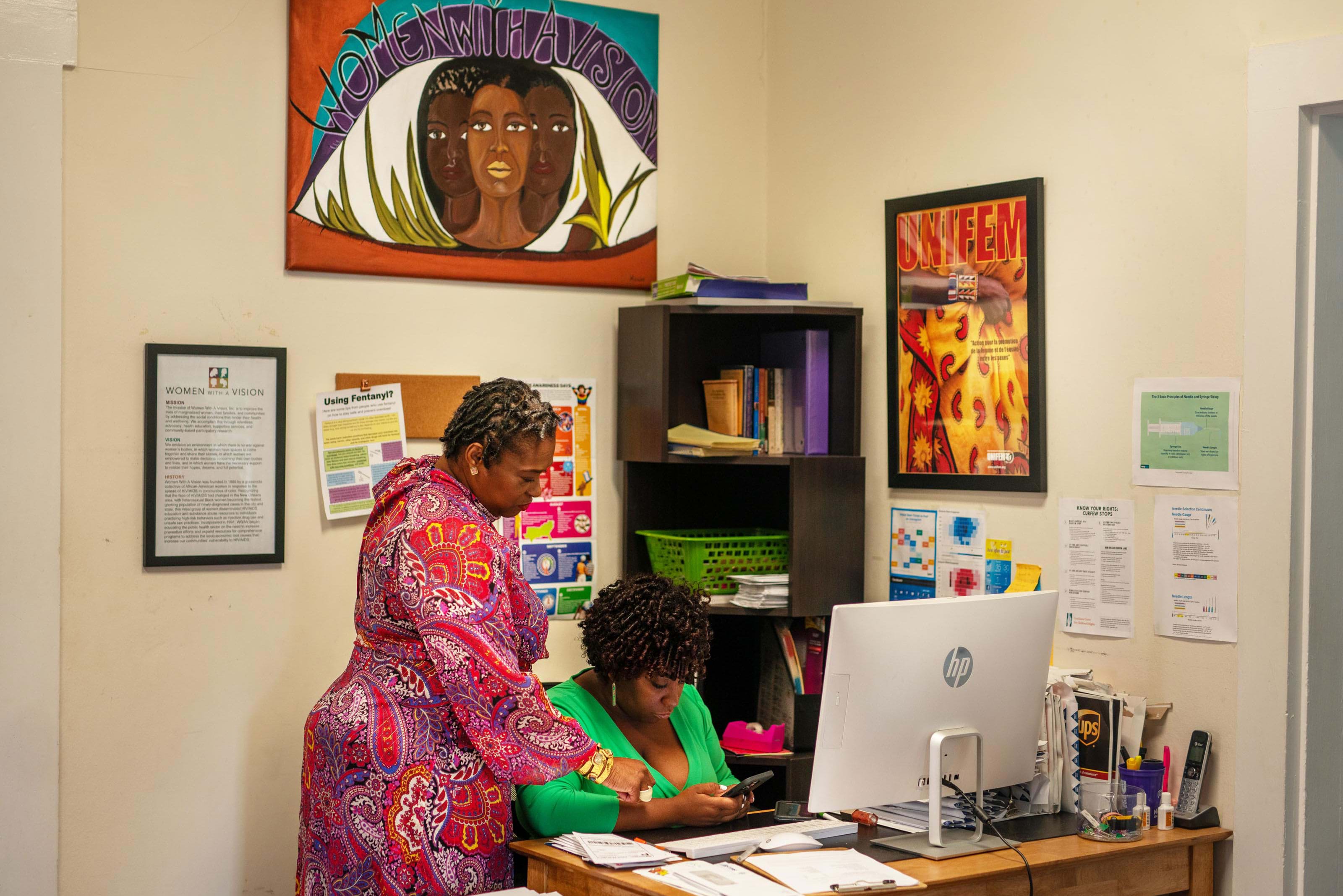 Deon looks over the shoulder of another woman in a bright green dress as they work on a crowded desk. The wall behind them is decorated with documents, books, and artwork. 
