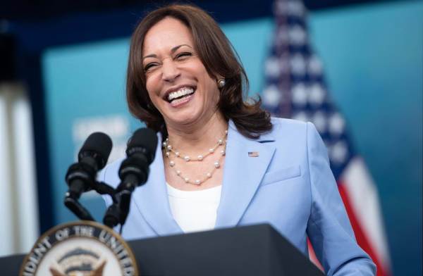 Vice President Kamal Harris is wearing a light blue blazer and white top and a pearl necklace, standing at a podium with the American flag in the background.