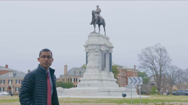 A man stands in front of a statue looking at the camera. He is wearing a blue puffer jacket, blue shirt, and red tie