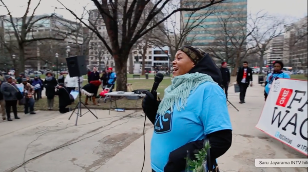 Photo of a Black organizer holding a microphone and smiling. There is a crowd and an obscured sign that says "Fair Wages"