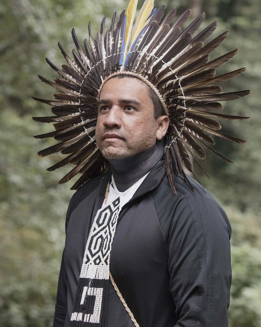 An Indigenous Brazilian man wearing a traditional feather headdress and bead necklace over a black long sleeve shirt in front of trees.