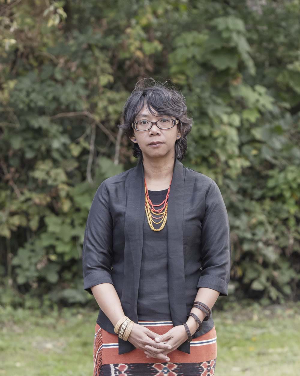 An Indigenous Dayak woman with short black hair and eyeglasses wearing a black jacket with a t-shirt underneath and a traditional orange Dayak skirt with florals and vertical lines. Behind her are green trees.