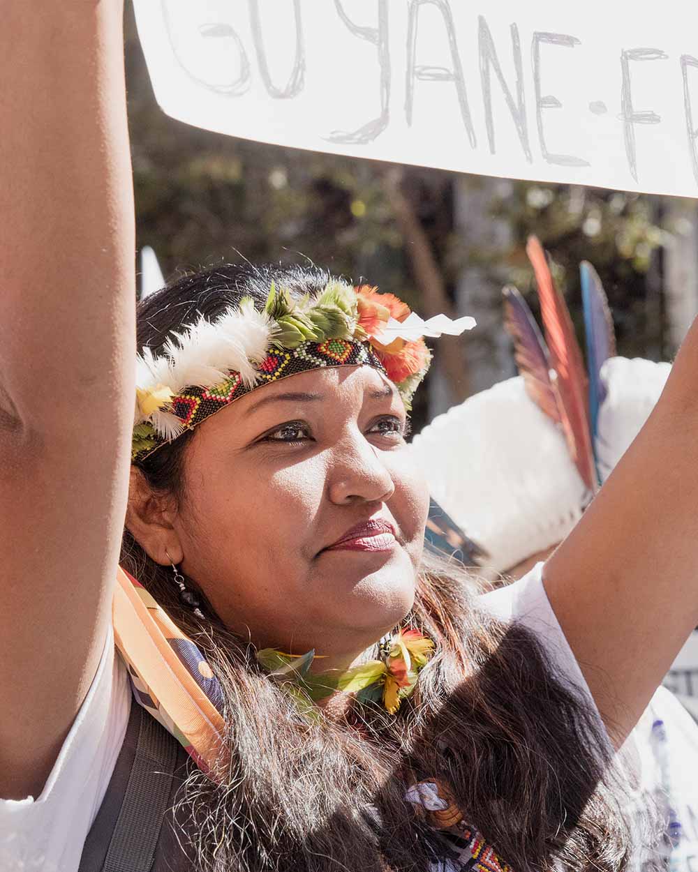 An Indigenous woman with long black hair and a white t-shirt wearing a feather headdress holding a sign at a protest.