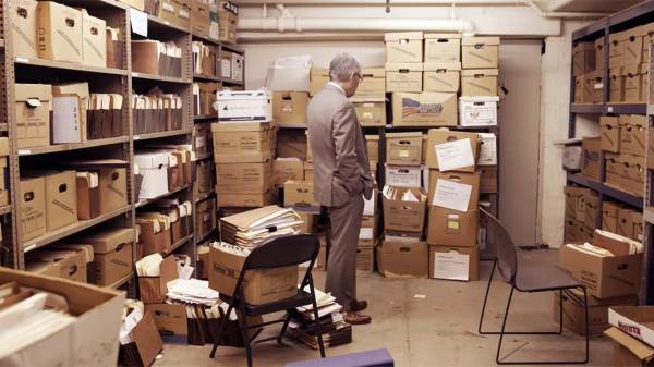 A man in a grey suit is staring at a stack of filing boxes.