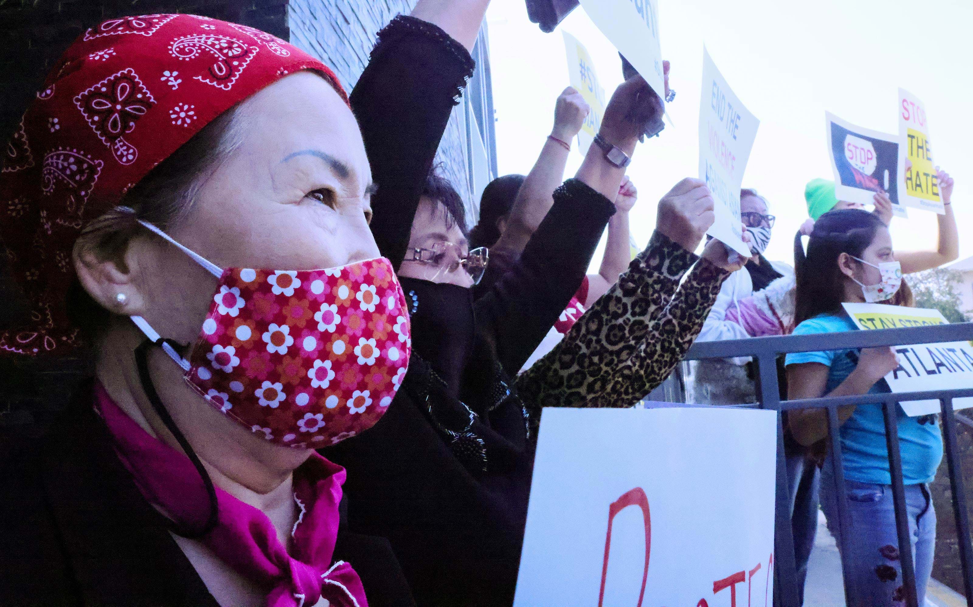 People holding signs protest hate crimes against Asian-American and Pacific Islander individuals. An individual wearing a red bandana and red mask holds a white sign and is in the foreground.