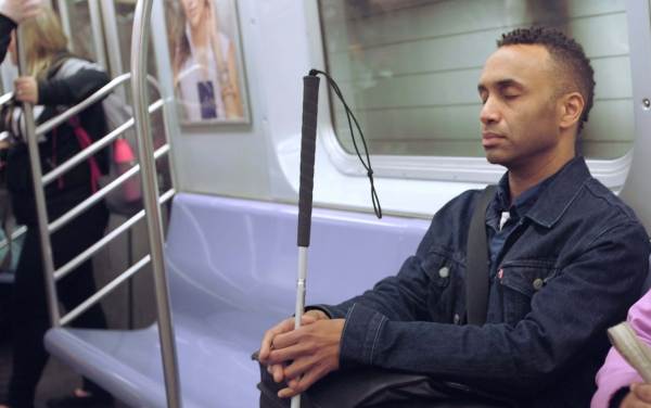 Black man wearing a blue jacket sits on a New York City subway train. He is holding a walking guide stick. 