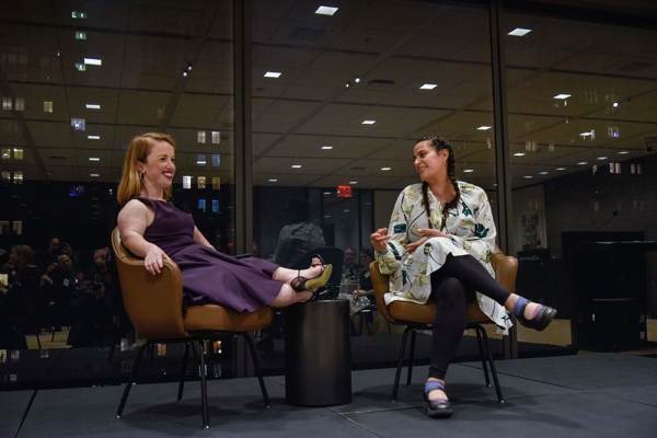 Rebecca, a white woman who is a little person with red hair, sits on a chair talking to Maysoon, another woman in a chair with two long braids and dark hair.