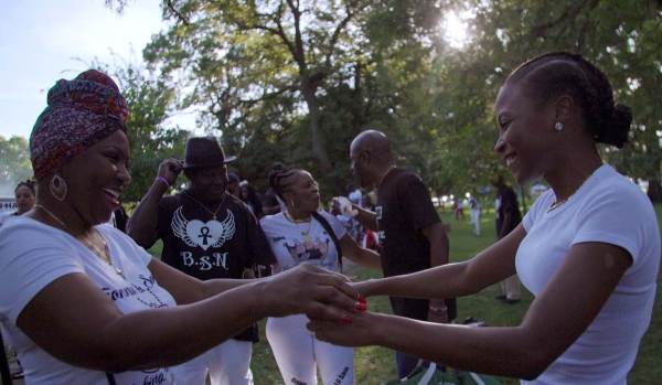 A photograph of two Black women smiling and holding hands while others gather in the background