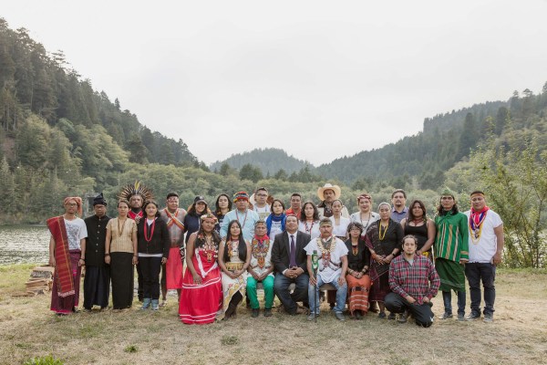 Group shot of Indigenous leaders from around the globe standing & sitting on a river bank in a wooded area.