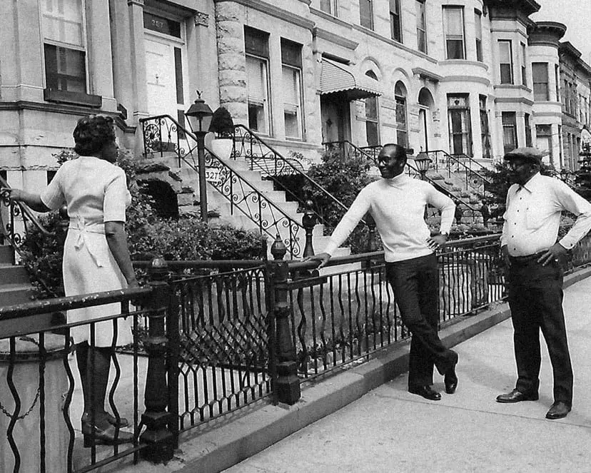 A black and white photo of Franklin Thomas talking to two residents on a brownstone lined black in Brooklyn.