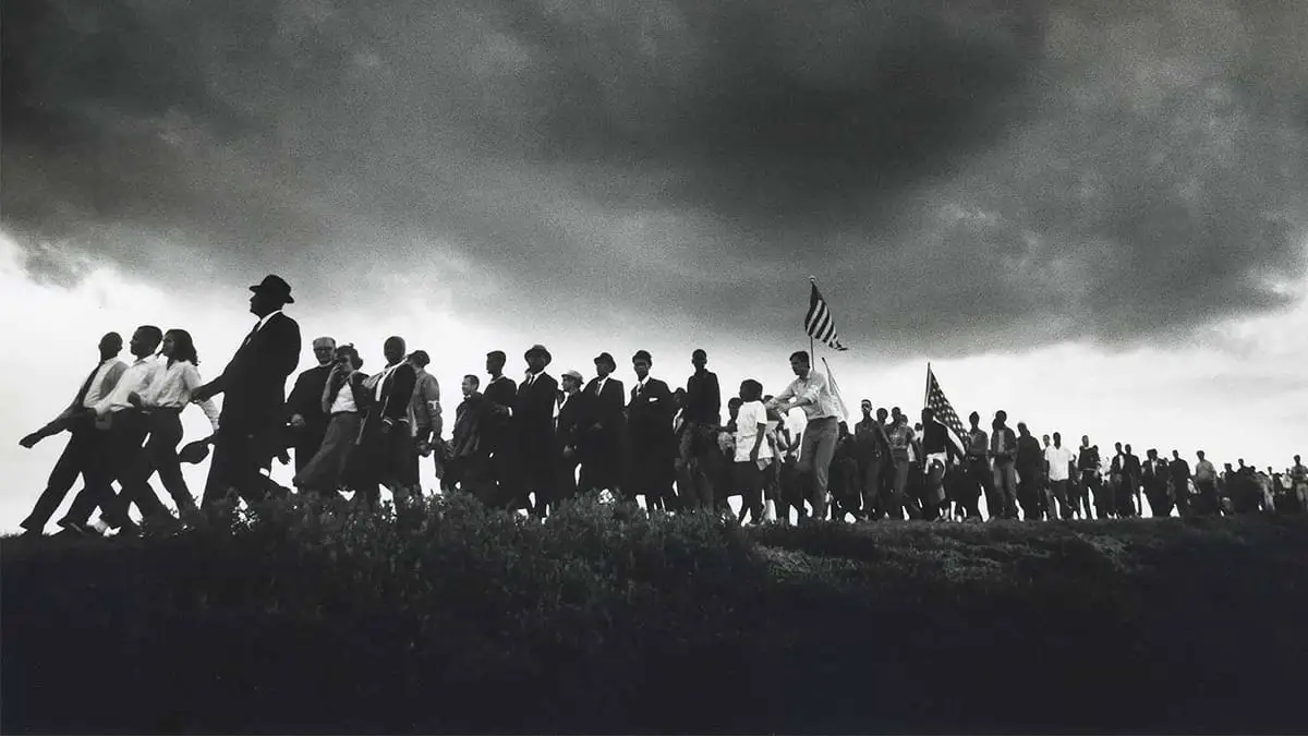 A historical photo of a crowd marching on a hill with a cloudy sky above them.