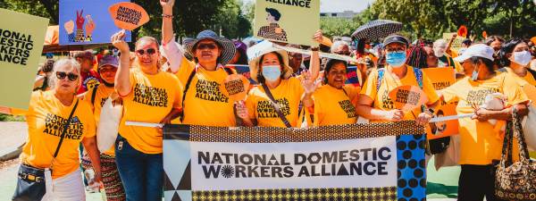 On a sunny day, demonstrators from National Domestic Workers Alliance hold signs and a banner with the organization’s name in bold font and colorful patterns. They’re are smiling and wearing bright gold t-shirts.