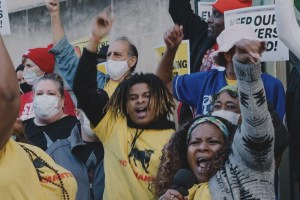 A crowd of people with raised fists and open mouths, many wearing masks, appear to be protesting or cheering. A man in a yellow shirt with dreadlocks and a woman in the foreground with a microphone are prominent.