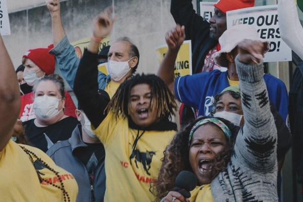 A crowd of people with raised fists and open mouths, many wearing masks, appear to be protesting or cheering. A man in a yellow shirt with dreadlocks and a woman in the foreground with a microphone are prominent.