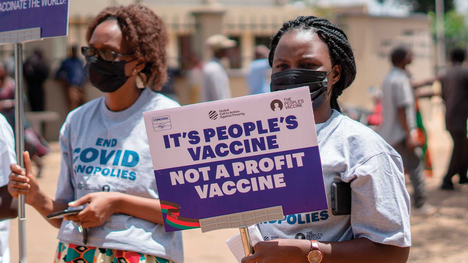 A Black woman wearing a medical mask holds a sign that reads “It’s people’s vaccine. Not a profit vaccine.” She’s surrounded by other demonstrators on a sunny day.