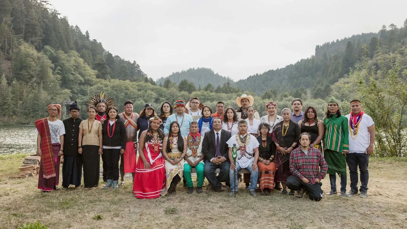 Against a sweeping backdrop of mountains and lush greens, two rows of Indigenous leaders wearing traditional headdresses and attire look on.