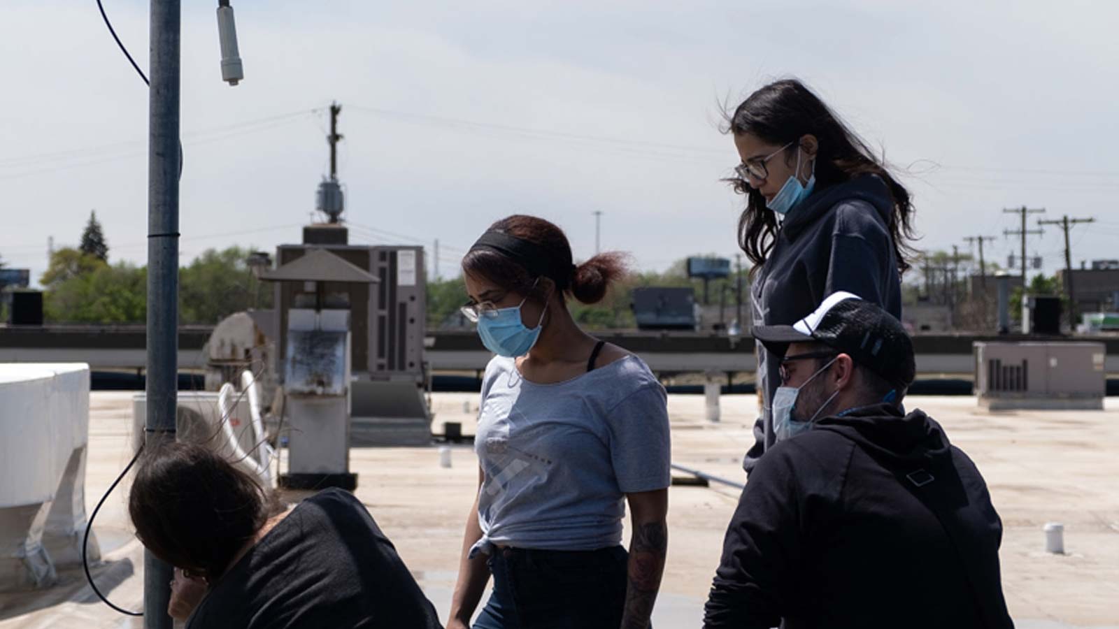 Four technicians install an internet receiver on a rooftop. They are all wearing medical masks and watching one worker making adjustments to a metal pole.