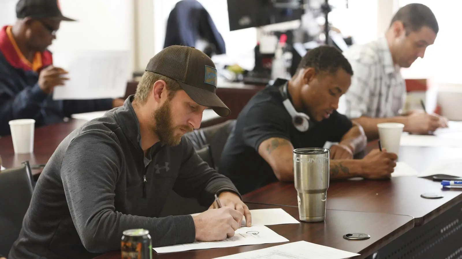 A man wearing a baseball cap fills out a document on a table. Behind him, other people are also filling out forms as light streams in from a window. 