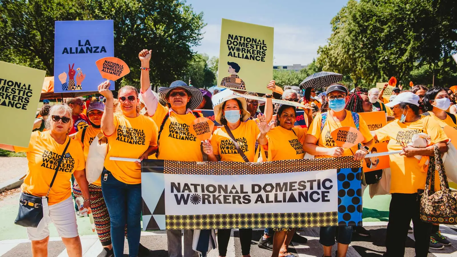 On a sunny day, demonstrators from National Domestic Workers Alliance hold signs and a banner with the organization’s name in bold font and colorful patterns. They’re smiling and wearing bright gold t-shirts. 