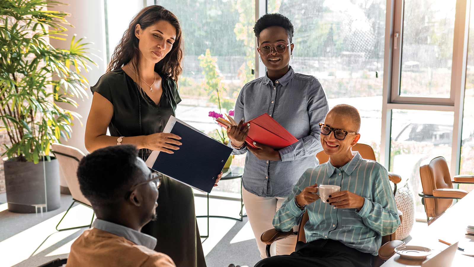 In a modern light-filled conference room, diverse office workers share a smile while a colleague sits in a wheelchair. Spread out on a nearby table are laptops and office supplies.