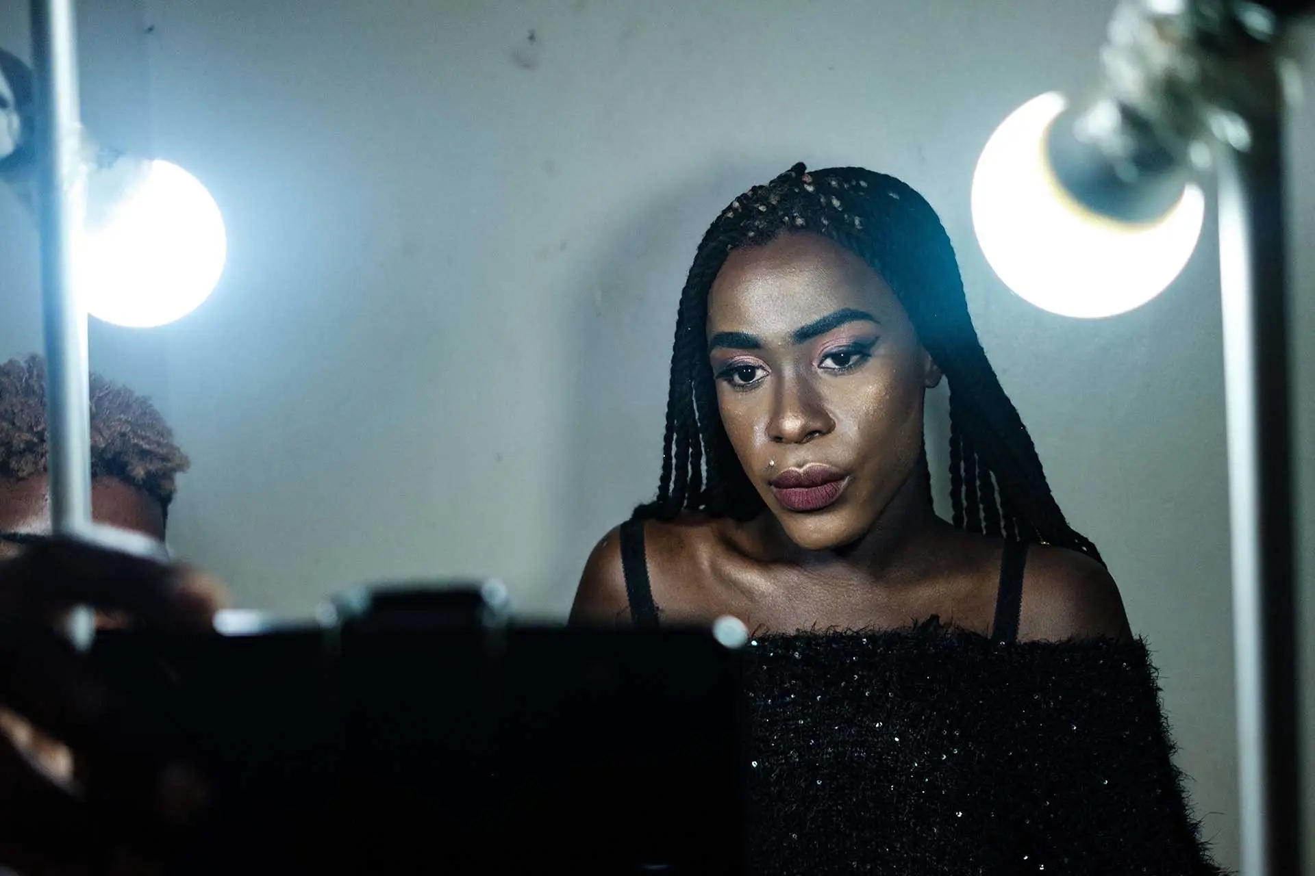 A Black woman in a glittery black top sits in front of a camera and lightbulbs, a blank wall behind her.