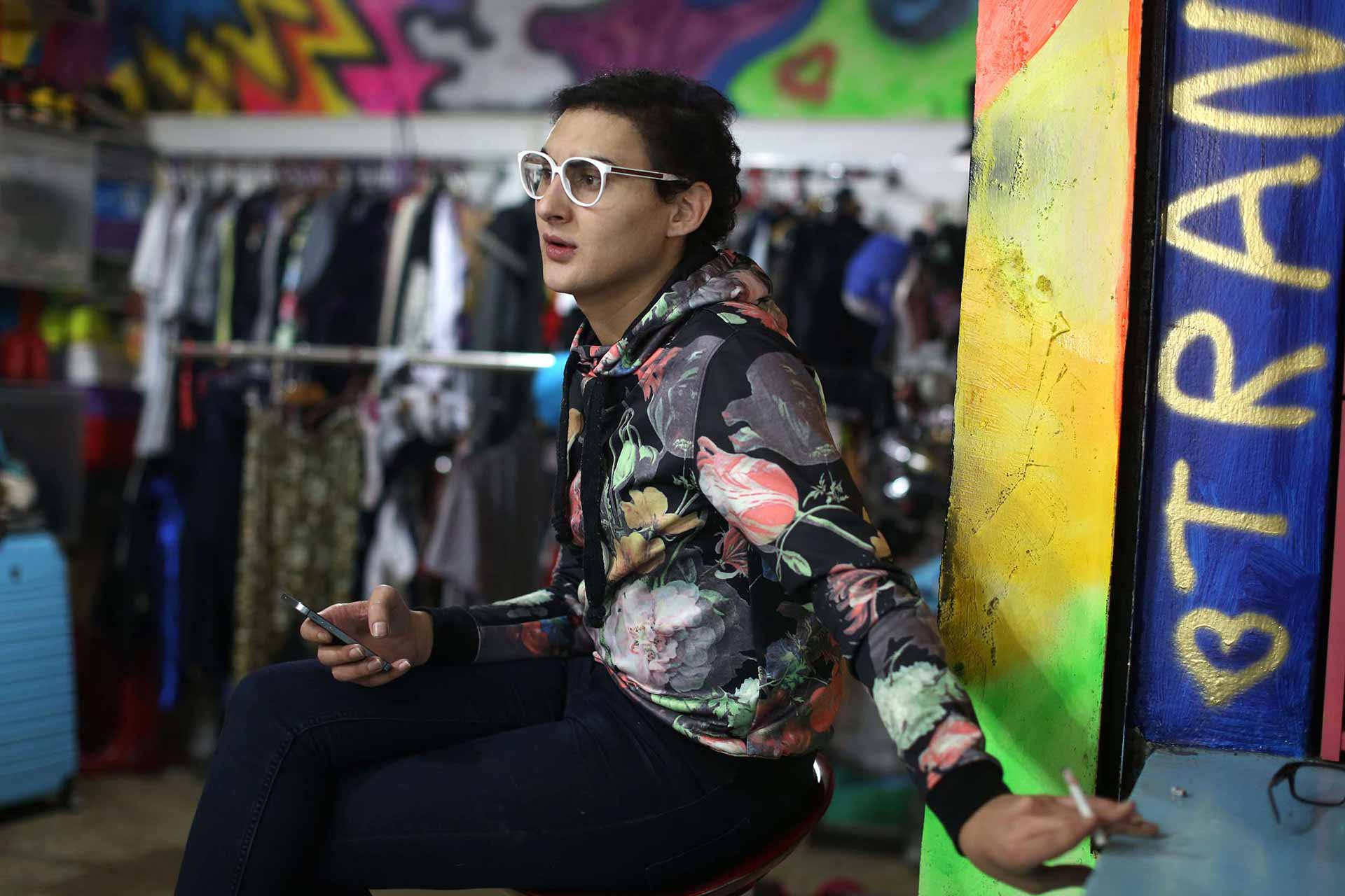 A fashion designer wearing a colorful print jacket and white glasses sits in front of racks of clothes. “Tran” and a heart are spray-painted in gold on the wall next to him.