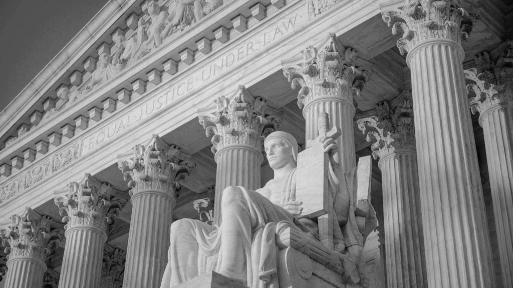 Back and white image of the exterior facade of Supreme Court of the United States. A statue of a seated figure holding up a stone tablet faces the sun.
