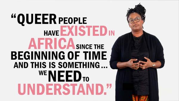 Selly Thiam has black dreads tied up in a knot and is wearing dark thick-rimmed eyeglasses. She has a nose ring and wears a black sweater over a black blouse and colored patterned slacks. She's standing next to copy that reads, "Queer people have existed in Africa since the beginning of time, and this is something we need to understand."