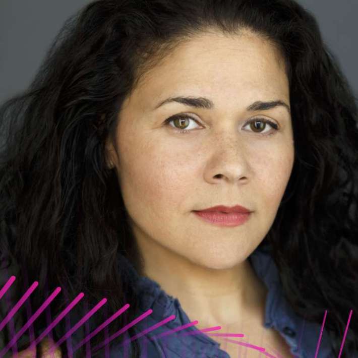 A light-skinned, biracial woman with dark, curly, shoulder-length hair and pink lipstick. She is wearing a blue, ruffled top and is in front of a grey backdrop.