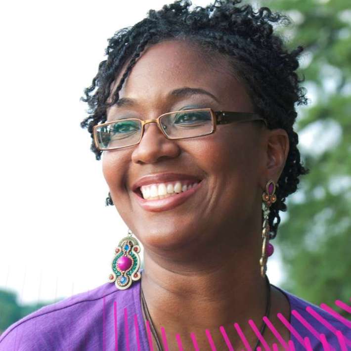 A Black, cisgender woman with brown skin and hair styled in two-strand twists. She is smiling and wearing makeup, glasses, dangling earrings decorated with jewel-toned stones, a necklace, and a V-neck purple dress. She is in a park and there are trees behind her.