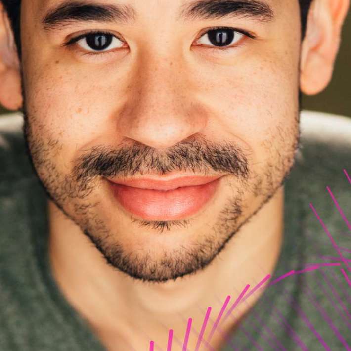 A multiracial man. He has dark brown, curly hair, wears a gray t-shirt, and smiles warmly at the camera.