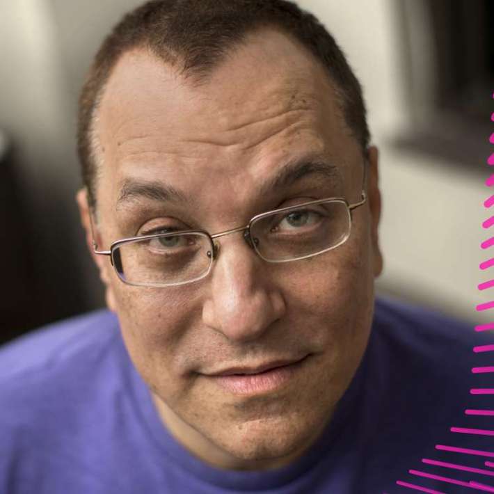 A white man with short brown hair and rectangular glasses. He wears a bright purple shirt and glances up towards the camera.