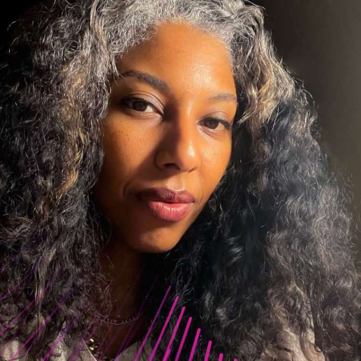 A brown-skinned woman in her 40s with brown eyes, curly salt-and-pepper hair, a white silk blouse, and a gold necklace. She is smiling and looking at the camera. The background is dark on the left side, lighter toward the right.