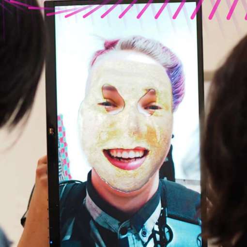 Two people viewing a screen. On the screen is a person with blond, purple, and pink hair wearing a digital mask and smiling brightly. A label reads, ""Masking Machine, M. Eifler."""