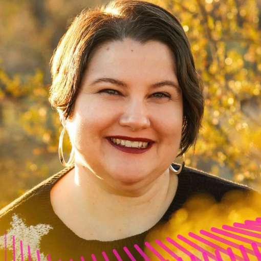 A light-skinned Mestizx with dark hair, silver hoop earrings, burgundy lipstick, and a black sweater with a white star. They sit in their scooter smiling and surrounded by golden creosote bushes.