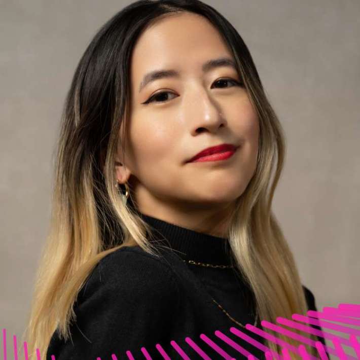 A Taiwanese American femme in her late 20s. She has dark hair with bleached blonde ends and wears red lipstick, a black turtleneck, and a small beaded necklace. She sits in front of a tan backdrop.