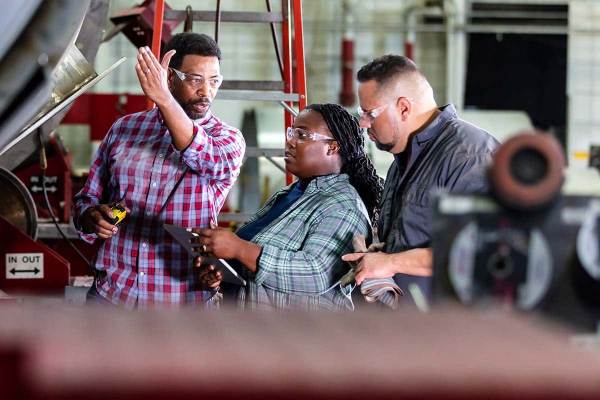 A man gestures toward a large machine on the factory floor. A woman and another man look in the same direction while taking notes. Around them is heavy machinery.