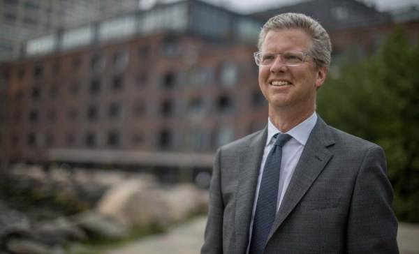Sean Donovan, wearing clear eyeglasses and a gray suit, looks on and smiles. Behind him are a large brick office building and a tree.