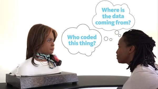 Stephanie Dinkins is a Black woman with black shoulder-length locs and wearing a white top. She's staring at a robotic torso of a black woman with shoulder-length brown hair. The word phrases "Who coded this thing?" and "Where is the data coming from?" appears in thought bubbles between them.