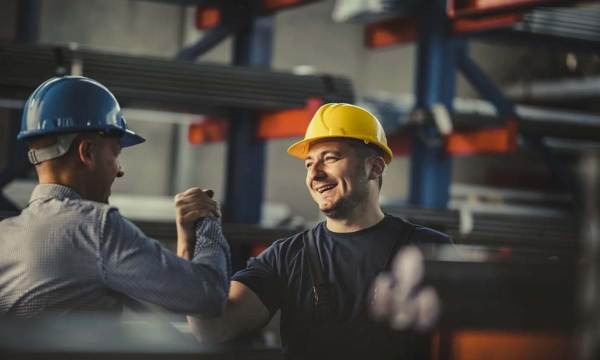 Two men wearing blue and yellow construction hats are doing a hand hug inside a warehouse.