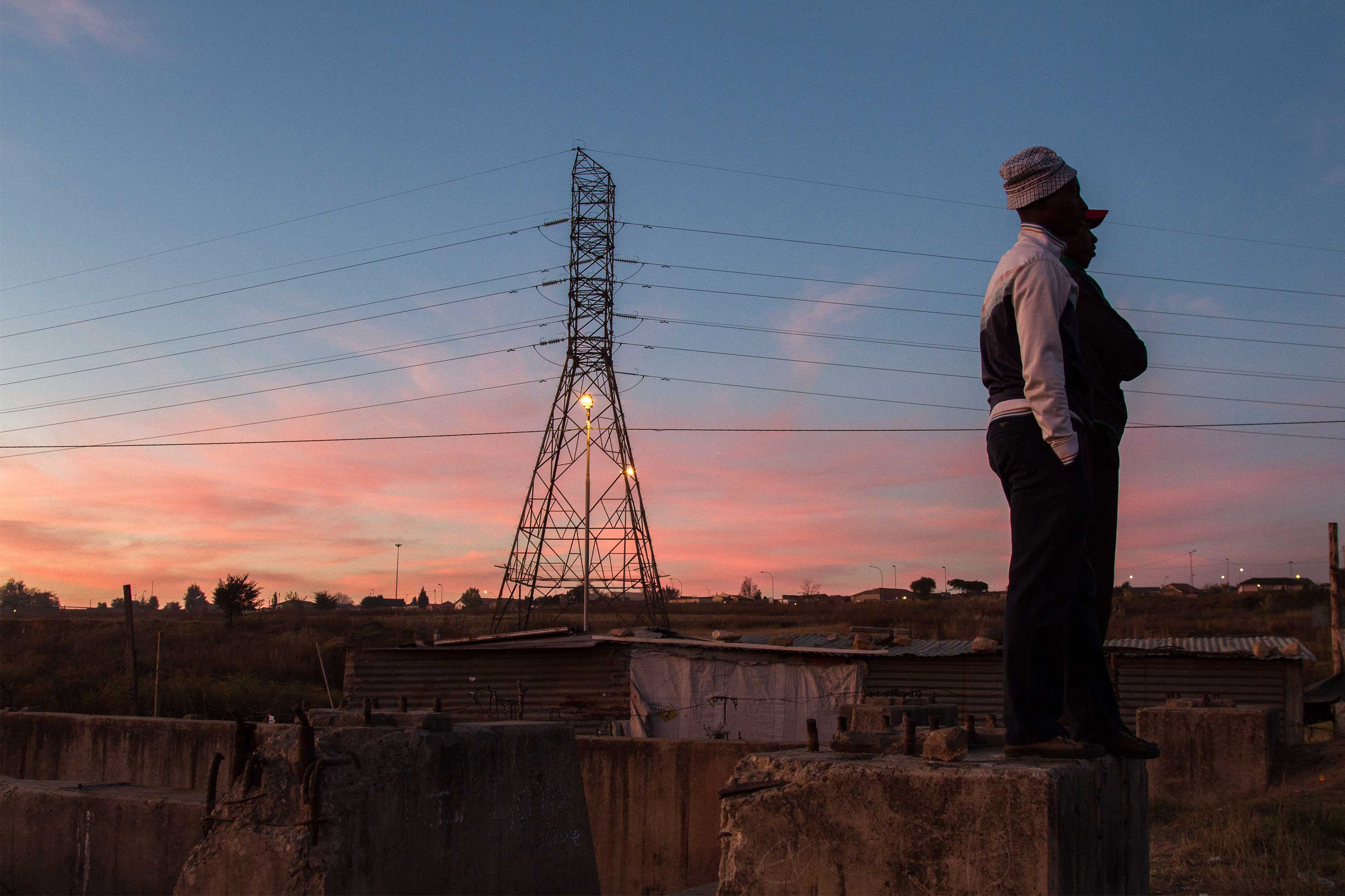 Two Black men look off in the distance. An electrical tower is in the background against a pink and blue sky.